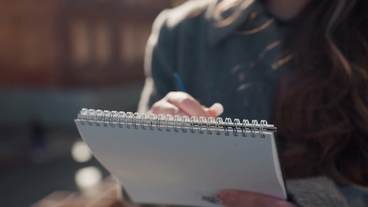 Pretty caucasian woman sketching notebook outdoors. Closeup hands holding spiral pad pencil, sunlit bokeh background, wavy brown hair over coat, focused expression, soft warm tones, paper texture