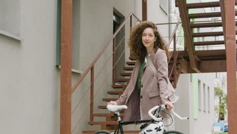 beautiful curly woman in formal clothes looking and smiling at the camera while standing with her bike outside in the city