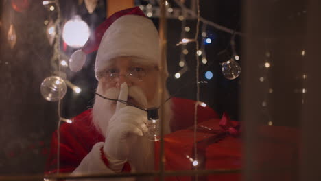 santa claus looks into the camera looking through the window and christmas lights. santa claus brought a gift