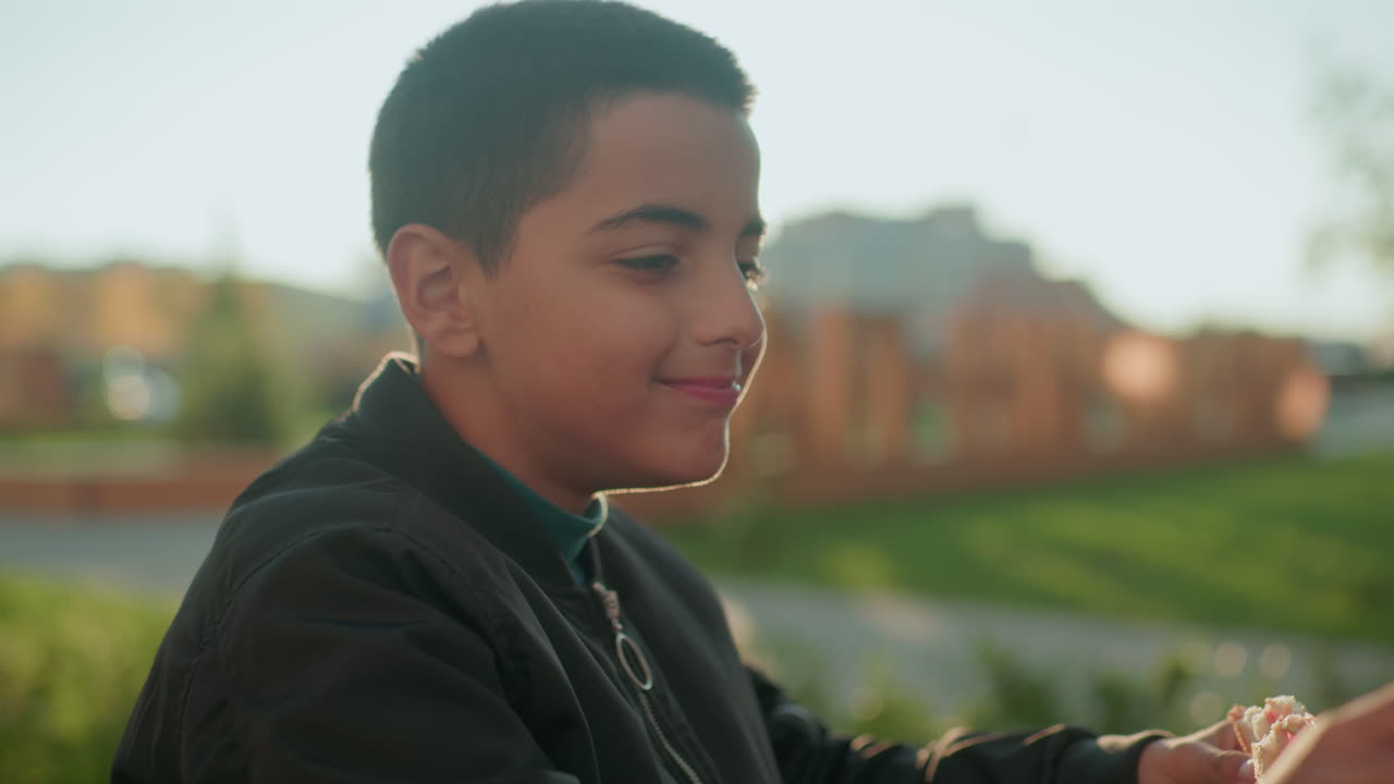 Boy drinking juice from glass bottle while holding sandwich outdoors at wooden table with warm sunlight in background, enjoying refreshing beverage and casual snack in relaxed urban atmosphere