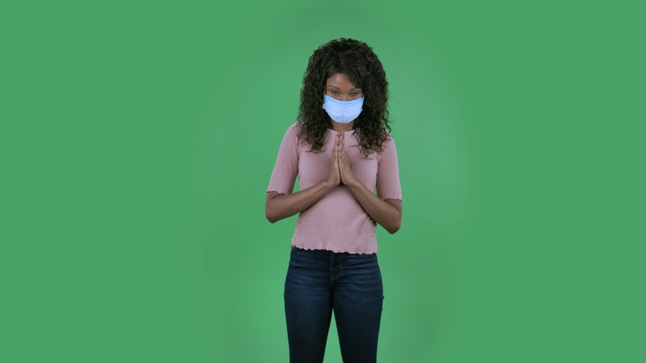 retrato de una hermosa mujer joven afroamericana con máscara médica está mirando directamente y reza con las manos juntas palma a palma. brunette ardiente con cabello ondulado en vaqueros y una blusa beige en una pantalla verde en el estudio. protección de la salud coronavirus
