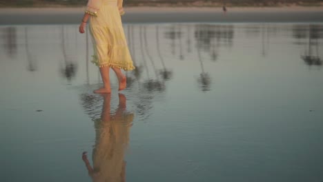 up close woman in a yellow dress calmly walking barefoot and swinging her arms at the shoreline at dawn