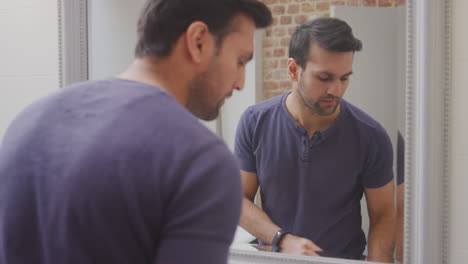 Hombre-Vestido-Con-Pijama-En-Casa-En-Un-Baño-Moderno-Usando-Productos-Para-El-Cuidado-De-La-Piel-Reflejados-En-El-Espejo