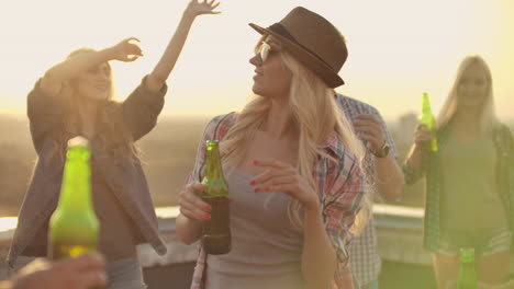 una joven con gafas y un sombrero se mueve en un baile con sus amigos en el techo. celebran una fiesta en el tejado con cerveza de botellas verdes de colores.