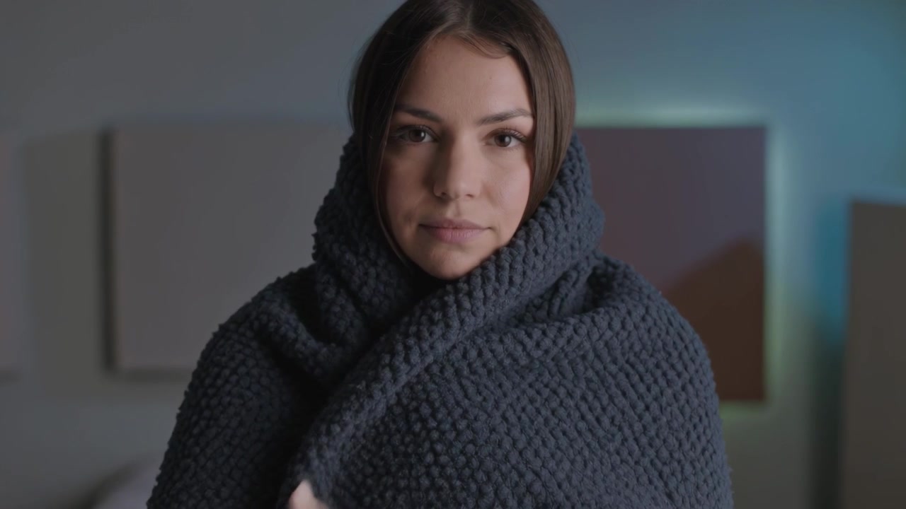 Sollevando una coperta grigia, una donna adulta si avvolge il busto in camera da letto, indossando una felpa leggera con un colletto.