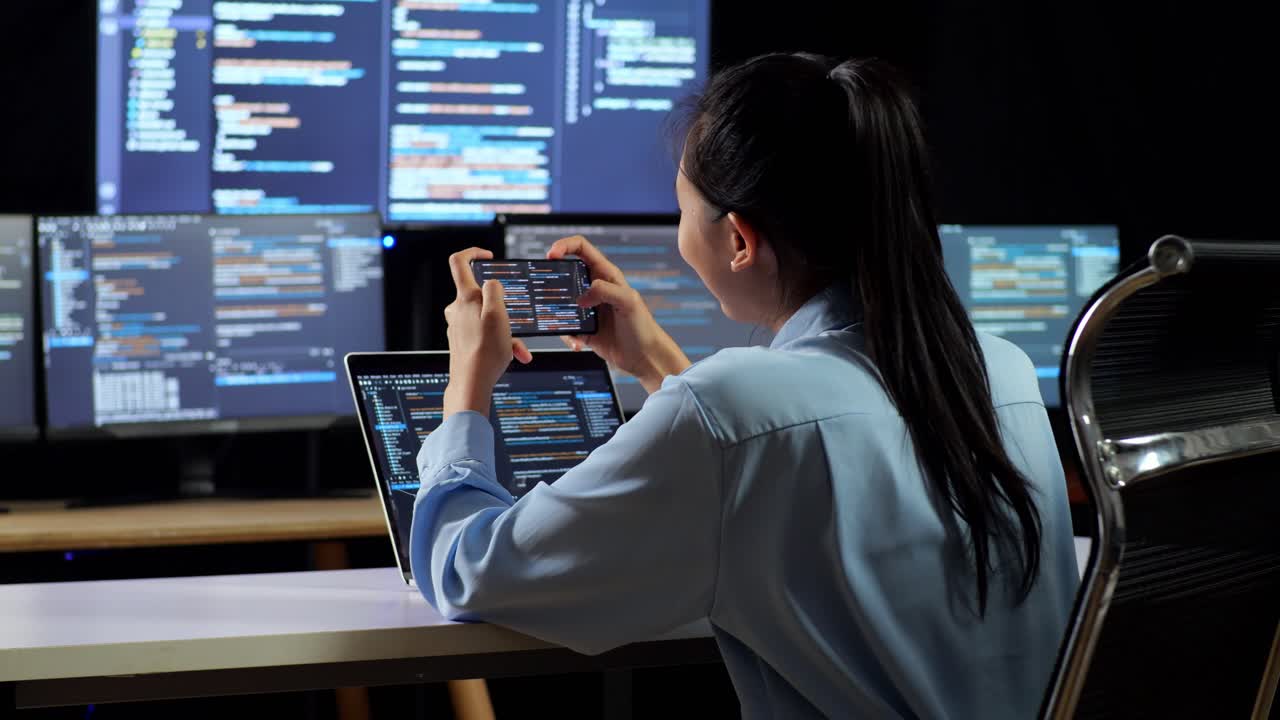 vista posterior de una programadora asiática mirando la base de datos en un teléfono inteligente mientras escribe código en una computadora portátil usando múltiples monitores que muestran la base de dados en los escritorios de la oficina
