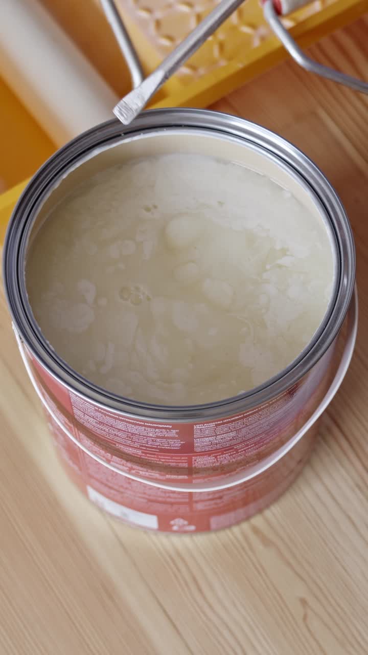 Top-down close-up of a person's hand prying open a metal lid of a white wood primer can using a flathead tool, then stirring with screwdriver, with paint tray and roller visible in the background