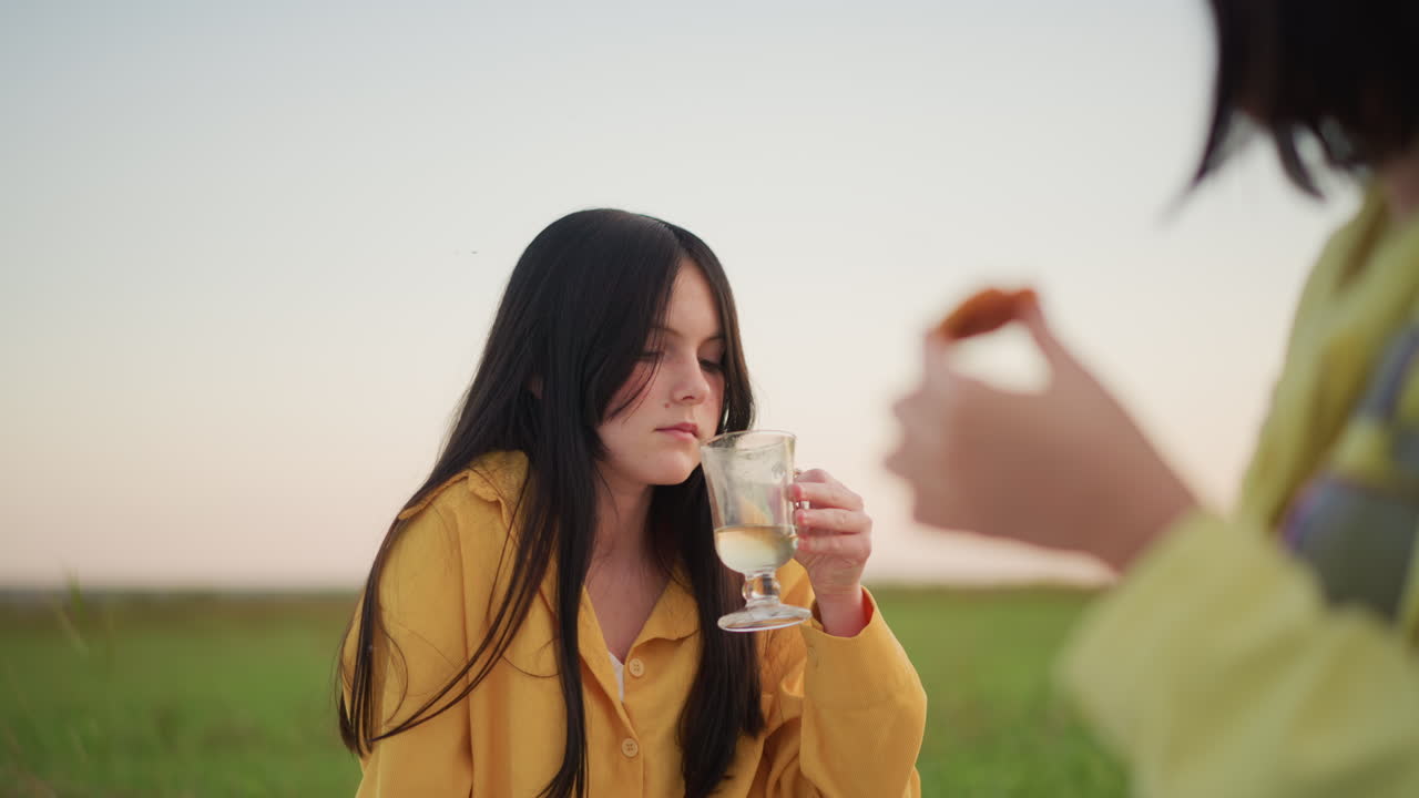 초원 거품 속에서 음료를 마시는 여성, 앞줄에 있는 친구는 쿠키를 들고 황금 시간대에 노란색 재킷을 입고 비눗방울을 불고 있어요. 유리 고블렛, 따뜻한 황혼, 솔직한 웃음, 부드러운 바람