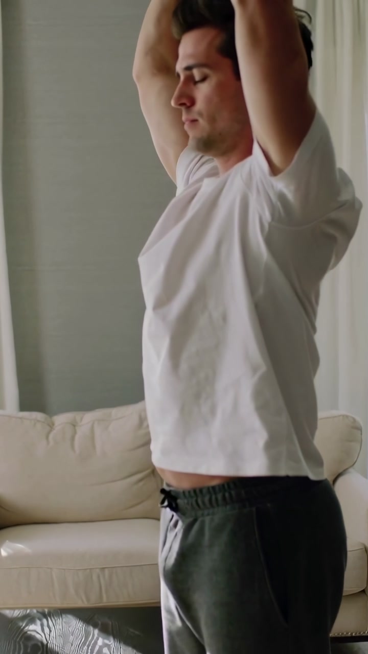Vertical video: Sunlight causing man in white tee gray sweats lifting arms up at home beside sofa