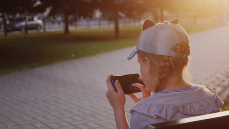 a girl plays on the smartphone sits in the park on a bench