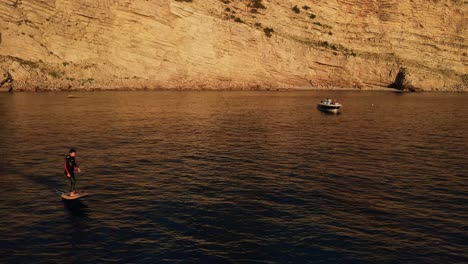 Aerial-follow-of-man-surfing-in-a-eFoil-electric-surfboard-at-Punta-Galera-in-Ibiza,-Spain-during-Sunset