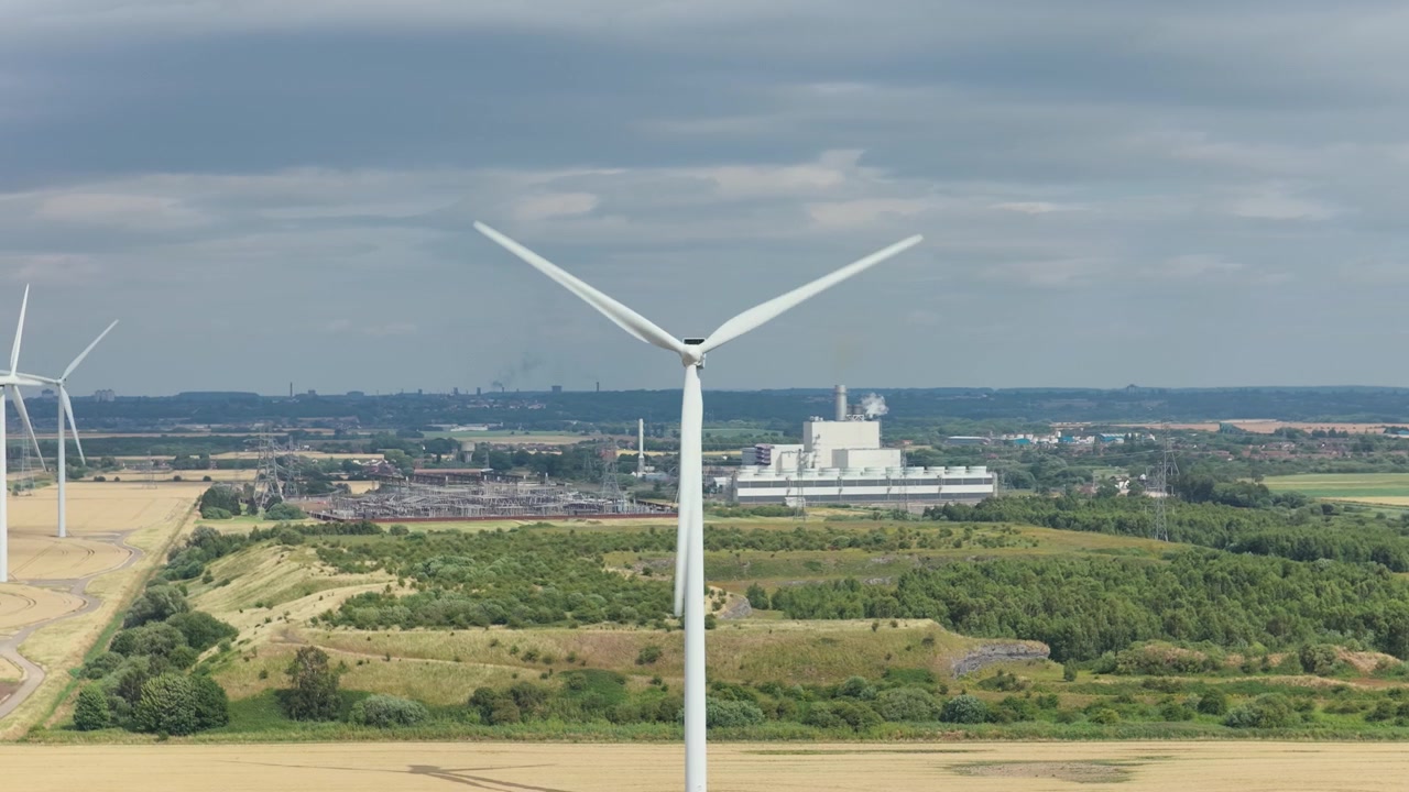 Multiple wind turbines rotate in golden fields, generating renewable energy. A significant industrial facility stands across green hills under a bright, cloudy sky.