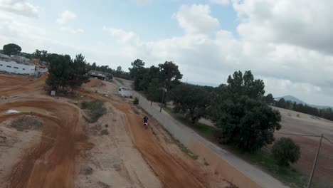 Epic-FPV-aerial-follow-of-motocross-rider-jumping-on-dirt-track,-cloudy-day