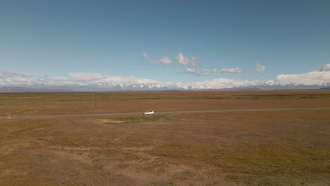 Silver-car-on-side-of-picturesque-road-within-dry-plains-with-snow-covered-mountains-in-backdrop