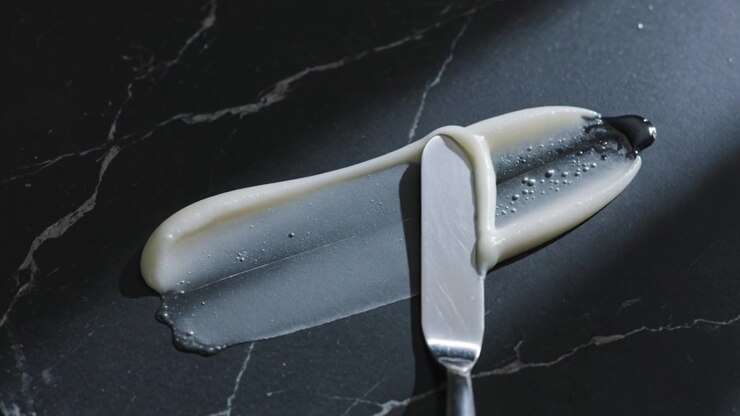 Spreading steel palette knife dragging pale cream across dark marble, smoothing, reducing droplets