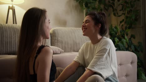 Two-young-women-having-good-time-together-in-apartment-near-sofa,-talking-and-hugging