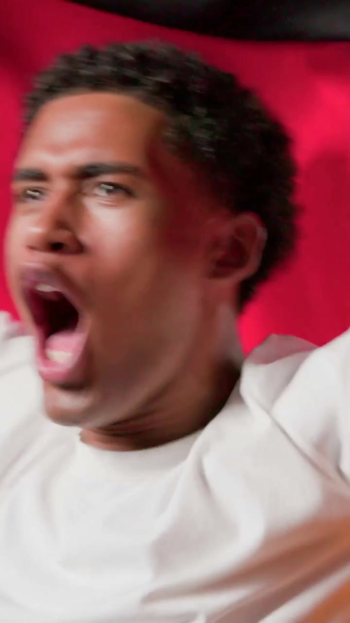 Vertical video: Shouting adult male in white tee, reacting to offscreen goal at red-black backdrop