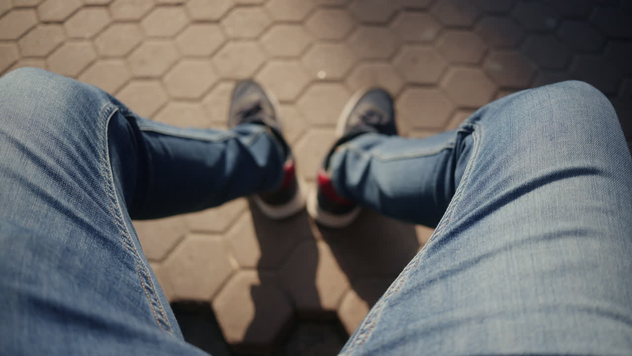 屋外の舗道に座っている男性の、ジーンズとスニーカーを履いた脚の俯瞰図。