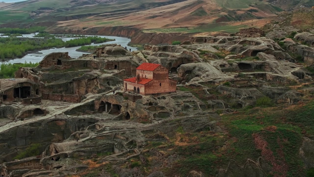 Majestátní let dronu nad jeskynním komplexem Uplistsikhe s výhledem na řeku Mtkvari. Filmové panorama drsné gruzínské krajiny a starověké historie při západu slunce.