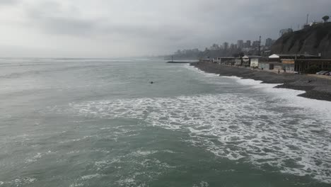 tiro aéreo frente a la playa en miraflores con el mar turquesa con olas tranquilas con gente en el agua, la playa con arenisca negra y diferentes edificios en un día nublado