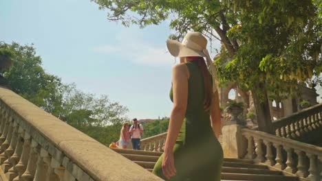 stunning footage of a girl in a green dress with hat walks up the stairs, joyfully observing cascada monumental-gaudí's fountain and parc de la ciutadella