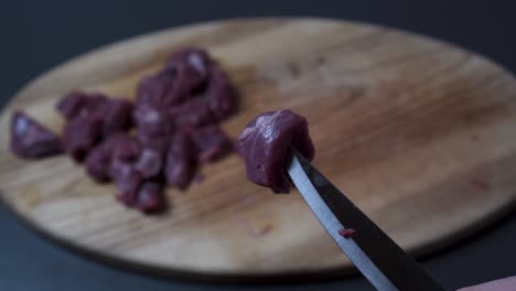 Small-Piece-Of-Raw-Beef-Impaled-On-A-Knife,-Wooden-Board-With-Sliced-Beef-In-Background---high-angle,-close-up