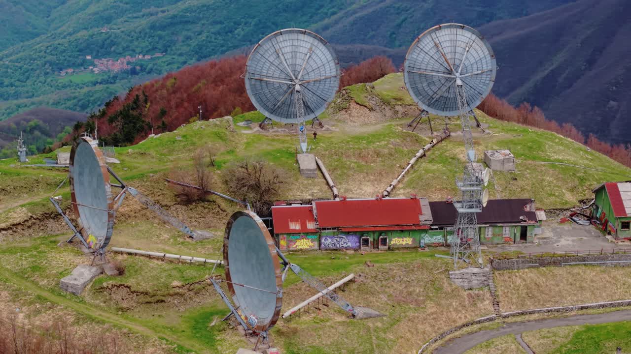 Old radar station with large satellite dishes on a grassy mountain ridge, aerial view