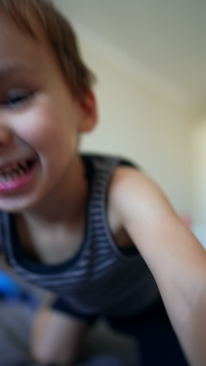 Smiling toddler sitting on the big bed plays with pyramid. Caucasian boy takes a toy detail and pushes it to camera. Vertical video.