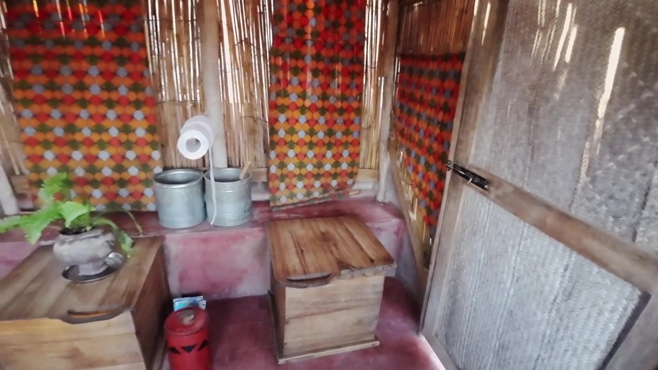 Ampia toilette compostante esterna con pareti in bambù, tende colorate, un tetto coperto e un lato aperto che si affaccia su una vista tropicale panoramica.