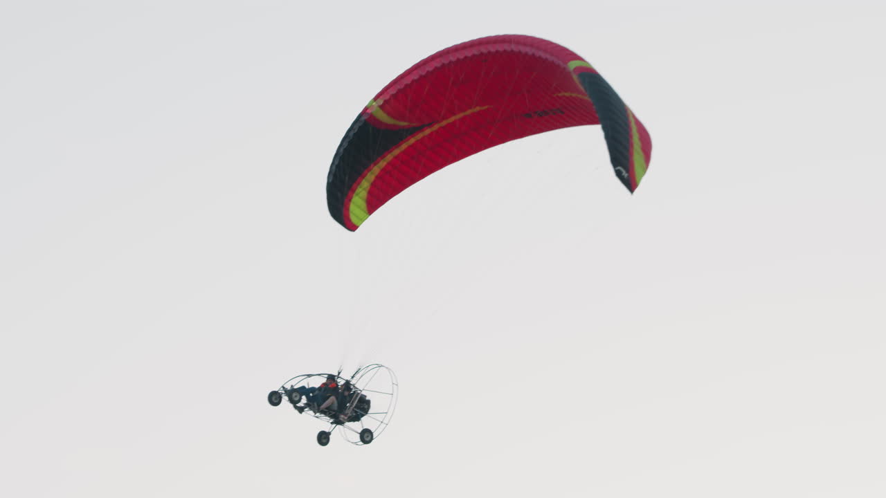Close up of two people seated in powered paraglider trike soaring under red and black canopy, engine cage and wheels visible against pale sky, lines taut as harnesses secure pilots