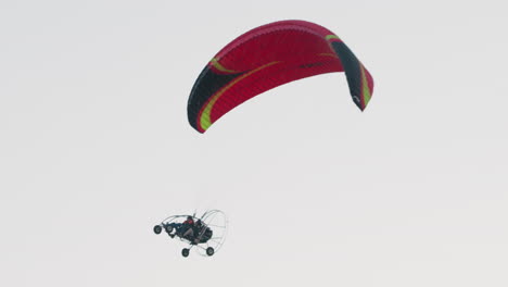 close up of two people seated in powered paraglider trike soaring under red and black canopy, engine cage and wheels visible against pale sky, lines taut as harnesses secure pilots