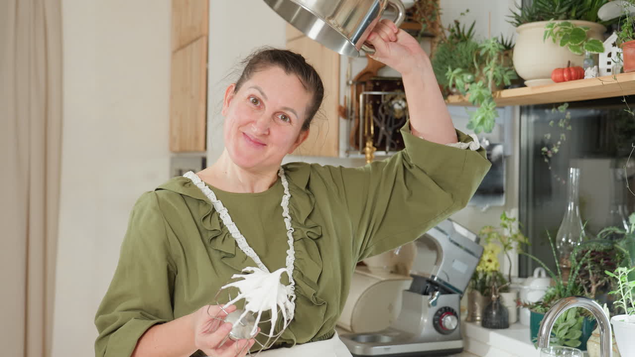 Chef in green apron playfully lifts empty mixing bowl upside down to demonstrate thick sticky dough clinging to electric mixer whisk while smiling in bright cozy kitchen