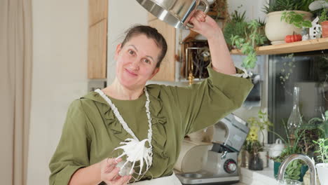 chef in green apron playfully lifts empty mixing bowl upside down to demonstrate thick sticky dough clinging to electric mixer whisk while smiling in bright cozy kitchen