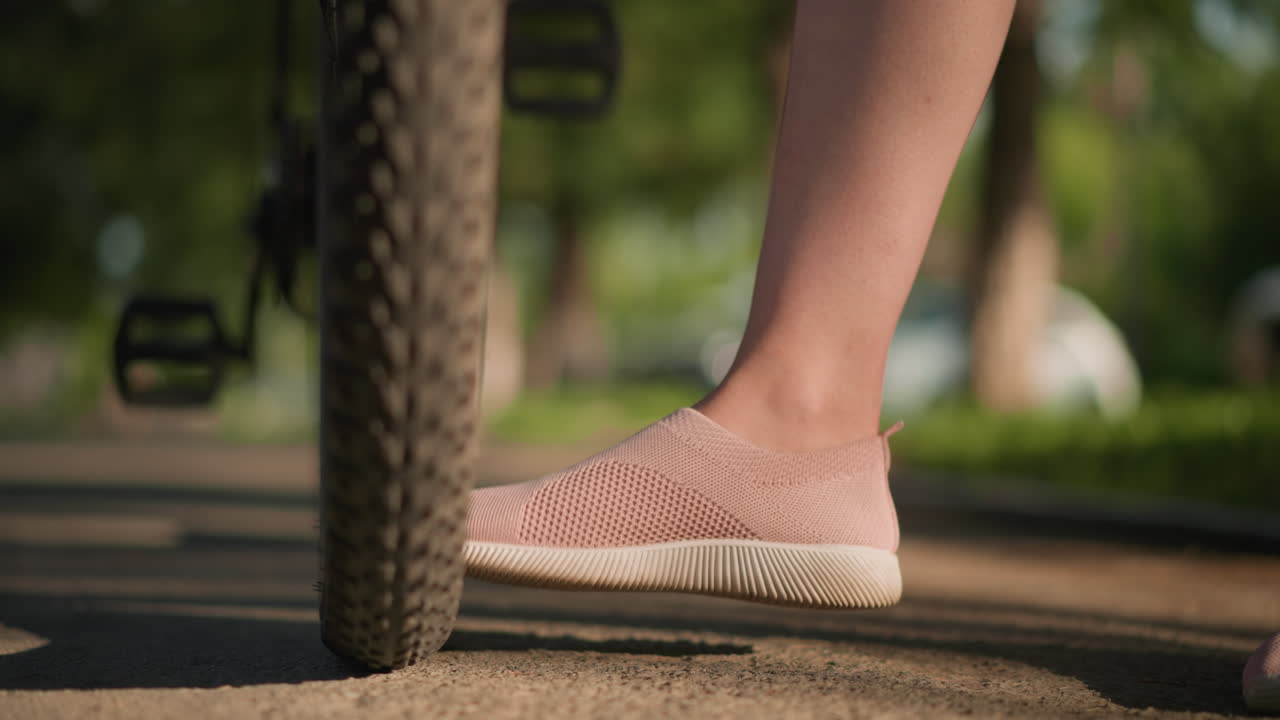 Close-up of legs wearing pink sneakers kicking bicycle tire to check air pressure, lush greenery and trees blur in the background