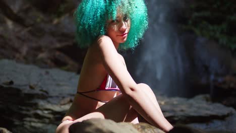 una chica de bikini de cabello verde se sienta en la base de una cascada que fluye en el caribe