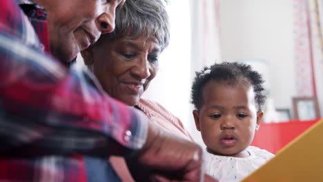 grandparents sitting on sofa reading book with baby granddaughter at home
