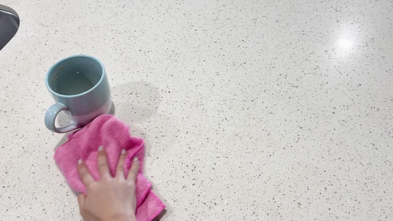 A person wipes a coffee spill with a pink cloth on a light countertop under bright lighting