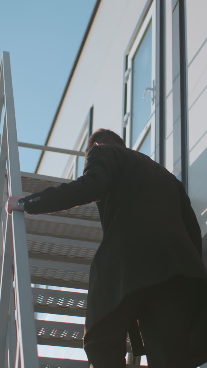 hombre de negocios en abrigo negro subiendo la escalera sosteniendo una barandilla de hierro y un maletín, con la luz del sol creando sombras dramáticas en la pared del edificio moderno a rayas y el reflejo de la cúpula en la ventana de vidrio
