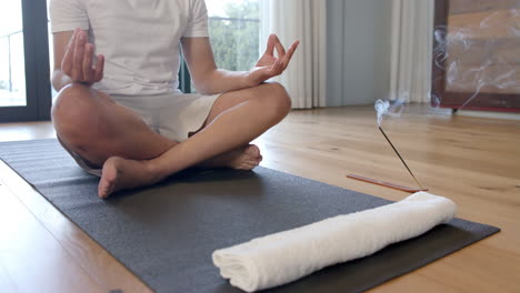 sección media de un hombre biracial haciendo yoga y meditando en casa, en cámara lenta
