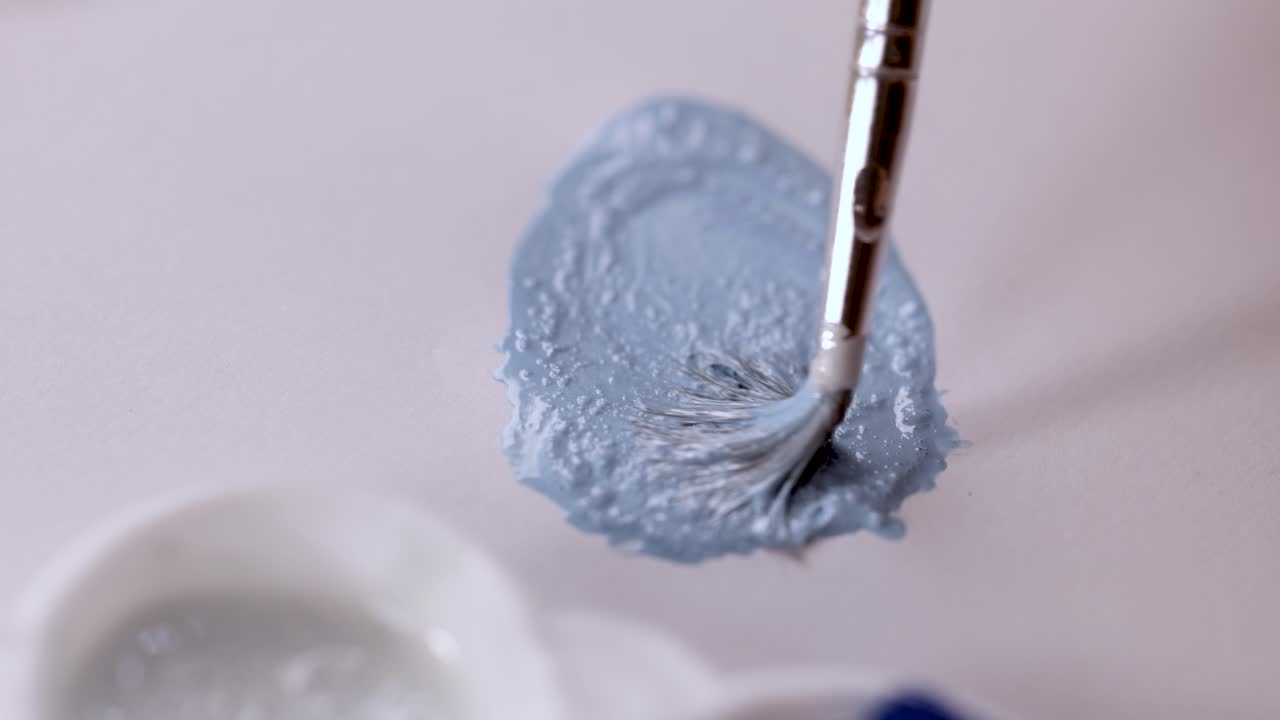Artist stirs light blue paint with brush on palette under soft, diffused lighting, macro perspective