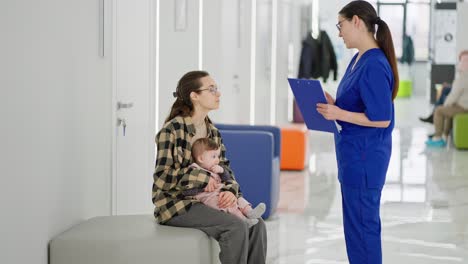 a young brunette girl the mother of a little baby girl sits in the corridor of a modern clinic and waits for her turn in the office of a pediatrician doctor. while a brunette girl doctor in a blue uniform comes up and reassures the young mother