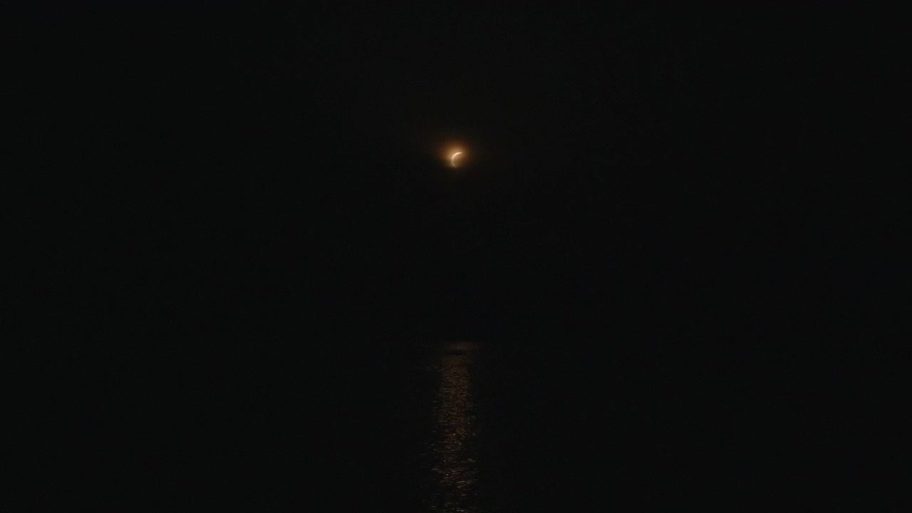 Piccola luna crescente brilla vicino al centro superiore sopra un oceano scuro, con un sottile riflesso ambrato che si estende sull'acqua immobile. La scena notturna rada appare tranquilla, remota, spaziosa e suggestiva