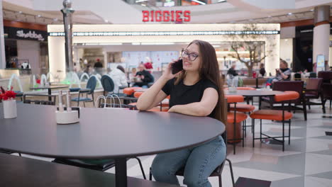 young woman happily making a phone call seated at a table in a busy mall, biggies sign visible overhead, with shoppers and diners around