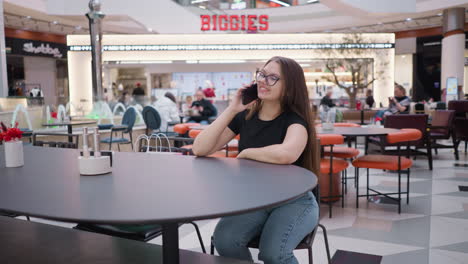 joven felizmente haciendo una llamada telefónica sentada en una mesa en un centro comercial ocupado, biggies señal visible por encima, con compradores y comensales alrededor