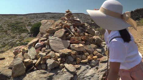 Female-in-summer-hiking-outfir-putting-stone-on-pile,-also-known-as-cairn,-in-arrid-landscape