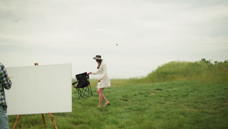 a woman in a white dress and hat is walking across a grassy field while carrying a chair. she approaches a painter and sets the chair in front of the painter's easel, preparing for an outdoor painting