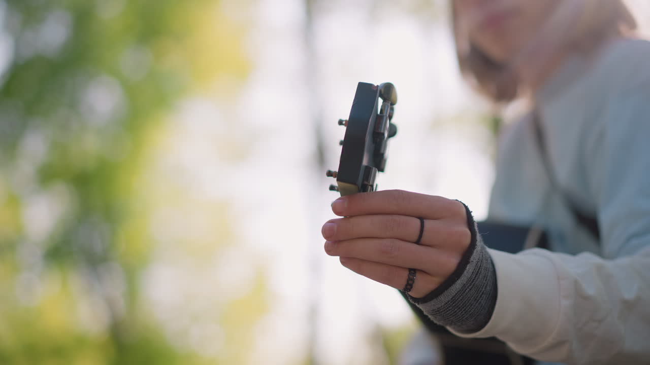 caucasian guitarist tuning acoustic guitar outdoors, busker adjusts headstock pegs, songwriter tests strings, student practices chords, denim sleeve and ring visible, bench under sunlight, soft bokeh