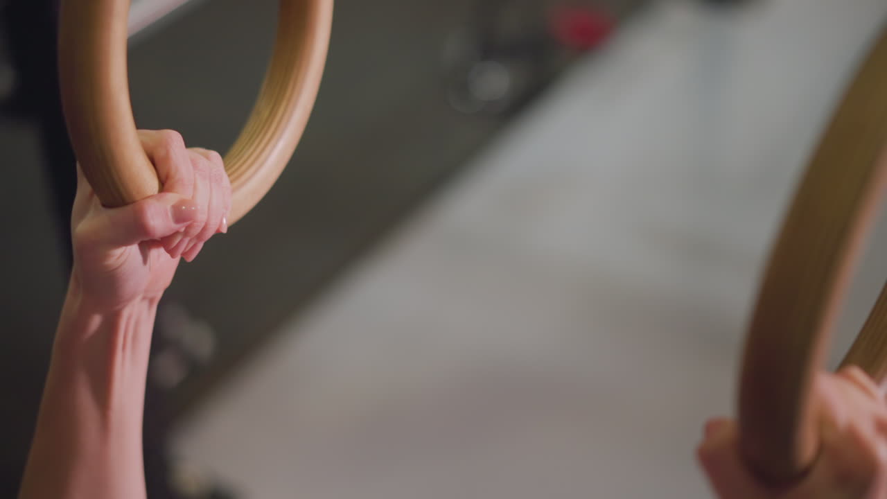 Asian woman gripping wooden gym rings, closeup study of hands and forearms during ring hold, sweat and callus detail reveal hard training, chalk dust and studio shadow create dramatic athletic mood