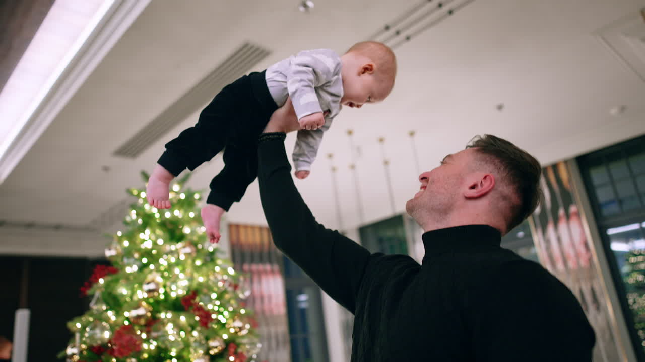 Strong muscular Caucasian man holding his son on the hand over his head. Christmas tree at backdrop. Low angle view.