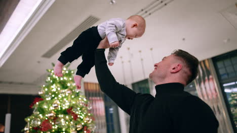strong muscular caucasian man holding his son on the hand over his head. christmas tree at backdrop. low angle view.
